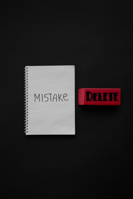 A white spiral-bound notepad with the word 'MISTAKE' handwritten in black ink is positioned on a dark background. To the right of the notepad, there is a red rubber stamp with the word 'DELETE' in bold black letters, also placed on the black surface. The arrangement is neat, with both items centered and clearly visible, emphasizing the concept of correcting or removing errors. The lighting is uniform, creating minimal shadows and highlighting the contrast between the white notepad, red stamp, and dark background. This scene visually reflects the idea of making careful decisions to avoid mistakes in processes such as rubbish removal or house clearance services, aligning with themes of efficient and accurate private waste handling as provided by companies like House Clearance Highbury. The simplicity and clarity of the objects support a professional and neutral tone suitable for service-related content, subtly underlining the importance of avoiding common pitfalls during independent collection or alternative waste disposal arrangements.