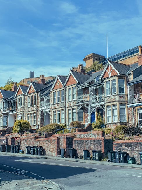 A row of Victorian-style terraced houses with ornate white ironwork and bay windows, situated on an elevated brick terrace with small landscaped gardens and low brick walls. The houses feature gabled roofs with slate tiles, brick and white rendered facades, and decorative elements typical of period architecture. In the foreground, there is a paved street with multiple black wheelie bins lined up along the curb, suggesting waste collection or disposal activity. The background shows modern office buildings with glass and brick facades, set against a partly cloudy blue sky. The scene indicates an urban residential area where private waste management services, such as rubbish removal, may supplement or replace communal council pickups, aligning with independent or on-site clearance options that companies like House Clearance Highbury could facilitate.