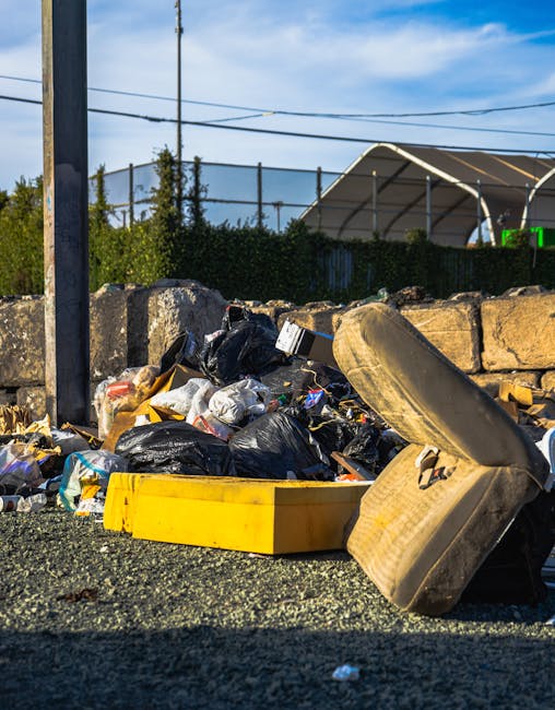 A collection of mixed waste and rubbish debris lies on the ground in a cluttered outdoor area, including black and clear plastic bags filled with waste, a yellow plastic container, a worn and dirty car seat or cushion partially resting on the ground with visible fabric and foam padding, and some loose paper and packaging materials scattered around. The waste is situated near a stone or concrete wall made of irregularly shaped blocks, with a metal pole visible on the left side of the image. In the background, there is a hedge or shrubbery, along with a large outdoor tent or industrial canopy structure with a curved roof, and overhead power lines against a partly cloudy sky. The scene suggests a location for either illegal dumping or a site awaiting collection, which could be relevant for private rubbish removal services offered by House Clearance Highbury, aligning with the context of alternative waste handling methods outside local authority disposal channels, all captured in natural daylight.