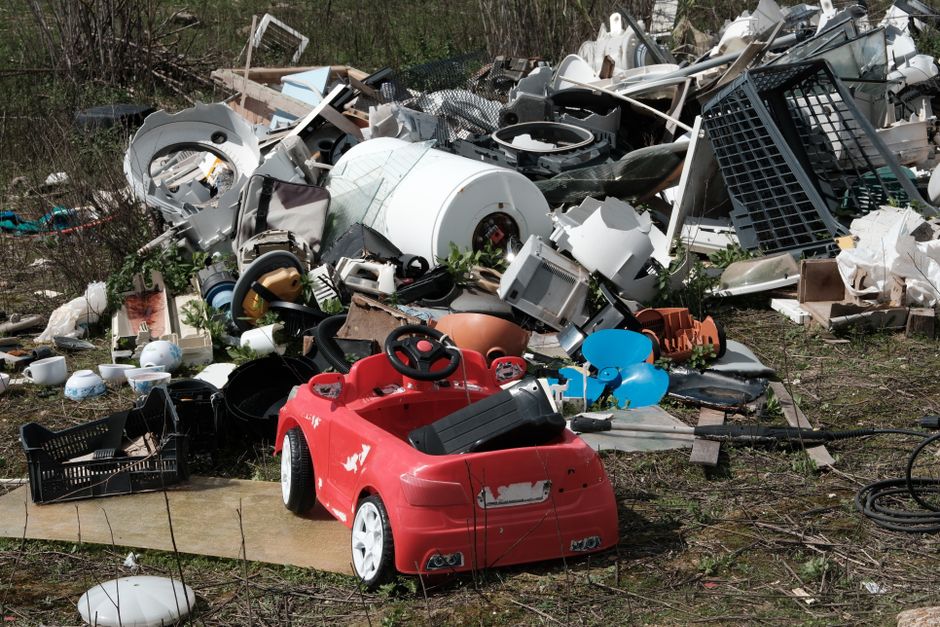 A large pile of assorted discarded household and electronic waste is scattered across an outdoor area, including a white washing machine lying on its side among various plastic objects, appliances, and broken items. In the foreground, a small red battery-powered toy car with minor paint wear is positioned on a patch of concrete beside some black plastic crates. The background shows more debris consisting of broken furniture, electronic components, and miscellaneous refuse, with patches of grass and bushes surrounding the area, indicating an unmanaged waste site. The scene displays a mixture of textures, from smooth plastic surfaces to rough, weathered materials, under natural daylight, highlighting the need for proper rubbish removal and waste disposal services, such as those offered by House Clearance Highbury, to address private waste clearance and prevent illegal dumping.