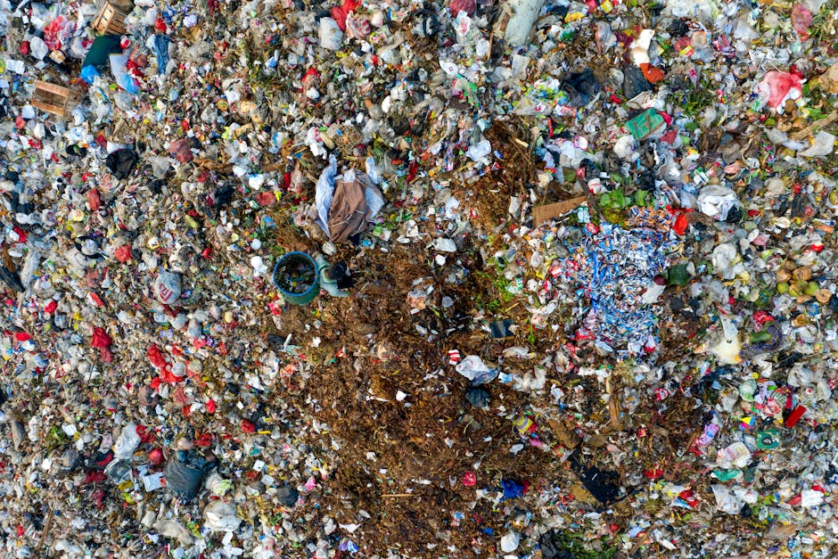 A collection of mixed waste and rubbish debris lies on the ground in a cluttered outdoor area, including black and clear plastic bags filled with waste, a yellow plastic container, a worn and dirty car seat or cushion partially resting on the ground with visible fabric and foam padding, and some loose paper and packaging materials scattered around. The waste is situated near a stone or concrete wall made of irregularly shaped blocks, with a metal pole visible on the left side of the image. In the background, there is a hedge or shrubbery, along with a large outdoor tent or industrial canopy structure with a curved roof, and overhead power lines against a partly cloudy sky. The scene suggests a location for either illegal dumping or a site awaiting collection, which could be relevant for private rubbish removal services offered by House Clearance Highbury, aligning with the context of alternative waste handling methods outside local authority disposal channels, all captured in natural daylight.