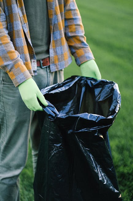 A person wearing a plaid shirt with shades of orange, grey, and blue, paired with grey pants and a brown belt, is standing outdoors on a grassy area. They are holding open a large black garbage bag with bright green gloves, preparing to dispose of waste. The background shows a well-maintained lawn with vibrant green grass, suggesting an outdoor setting such as a garden or park. The scene depicts an individual engaged in rubbish collection or waste disposal, which relates to independent waste handling or on-site clearance services sometimes utilized as an alternative to municipal rubbish collection. The focus is on the person’s hands and the open bag, emphasizing the task of rubbish removal, consistent with services offered by companies like House Clearance Highbury. The natural lighting indicates daytime, with no visible background structures or additional objects present, providing a clear view of the waste disposal action in progress.