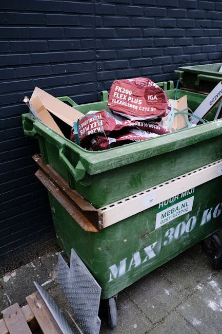 A large green wheelie bin filled with red and black bags of gravel or aggregate materials, positioned beside a dark brick wall on a pavement. The bin has a white label on the side and is supported on a wooden pallet, suggesting it is used for waste or rubble collection. The bags are partially open, with some spilling slightly over the edges of the bin. Surrounding elements include another bin of similar size and a small piece of metal grating or interior flooring leaning against the lower part of the bin, indicating a site of waste clearance or material disposal. The setting appears to be an outdoor area adjacent to a building or property, suitable for private rubbish removal or on-site debris clearance, with the scene capturing the practical use of a household or commercial waste collection service by House Clearance Highbury.