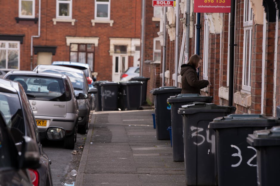 A residential street scene featuring a row of black wheelie bins placed along the sidewalk in front of red-brick terraced houses. The bins are lined up closely, some with lids slightly open, and have white spray-painted numbers on their fronts. A woman dressed in a dark coat is seen on the right side of the image, leaning over a bin while handling rubbish. Parked cars, including a grey hatchback and other vehicles, are positioned along the street, occupying the designated parking spaces adjacent to the sidewalk. The houses in the background have white-framed windows and brick facades, with some featuring small front gardens or steps leading to the doors. Overhead, a portion of a red 'Sold' sign is visible, attached to a building, suggesting recent property transaction or sale. The overall scene depicts a typical urban setting with waste management bins likely awaiting collection, reflecting local disposal and rubbish removal practices commonly found in residential areas. The lighting indicates daylight, and the environment appears clean apart from some minor litter on the pavement, aligning with the need for regular rubbish collection services. House Clearance Highbury provides waste removal solutions, including on-site clearance and private disposal options near this type of setting, where municipal collection might be bypassed or supplemented.