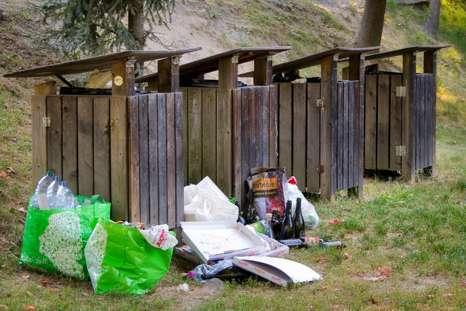 A row of four wooden outdoor latrine units with dark brown weathered panels and flat, overhanging roofs is situated on a grassy area, surrounded by trees and natural ground cover. In front of the units, there is a collection of scattered rubbish, including a green plastic bag filled with empty plastic bottles, a white pizza box with food residue, a black and orange bag of barbecue charcoal, several glass bottles, and various other litter items. The scene suggests an informal outdoor waste disposal area, potentially associated with private or unregulated rubbish handling, with the rubbish haphazardly piled near the structures amid a natural environment. The lighting indicates daytime, with soft natural light illuminating the scene, emphasizing the contrast between the wooden structures and the refuse in the foreground.
