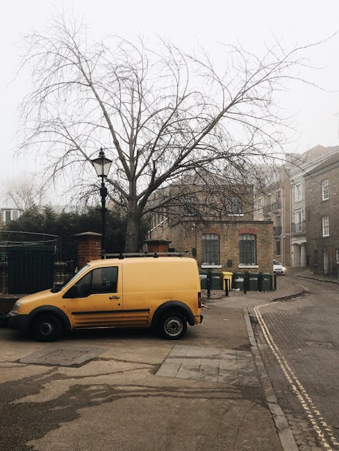 A yellow commercial van, typical of those used for private rubbish removal services, is parked on a paved street in front of a leafless tree with a textured bark surface. Behind the van, there is a row of green wheelie bins positioned along the curb, indicating waste collection activity. The background includes a small brick building with two windows, and a black wrought iron fence partially surrounds this area. To the right, a gentle street incline leads past residential buildings with multiple stories and light-colored facades, characteristic of urban housing. A vintage-style street lamp with a black metal post stands near the tree, contributing to the street scene's navigational environment. The overall atmosphere appears overcast with diffused daylight, and the setting suggests a typical outdoor rubbish collection or private waste disposal context, aligning with services offered by House Clearance Highbury and other independent waste handling providers.