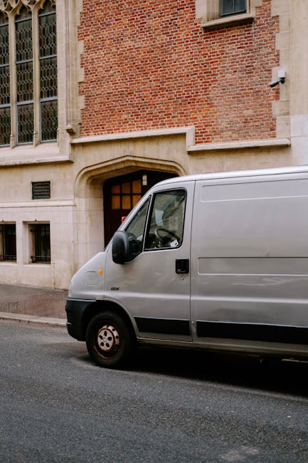 A silver cargo van is parked on a paved street in front of a red brick building with large window panes and stone framing around the entrance. The van is positioned on the right side of the image, with the driver's side facing the viewer. Its smooth metallic surface reflects natural daylight, and the black side mirror and wheel arch provide contrast against the light grey body. The building's facade features traditional architectural details, including stone window surrounds and a small security camera mounted near the top right corner. The environment appears clean and orderly, suggesting an urban setting where private or alternative waste collection methods, such as rubbish removal services from companies like House Clearance Highbury, might operate. The scene captures a typical street setting suitable for independent waste disposal or clearance work, with the van ready for loading or collection purposes.